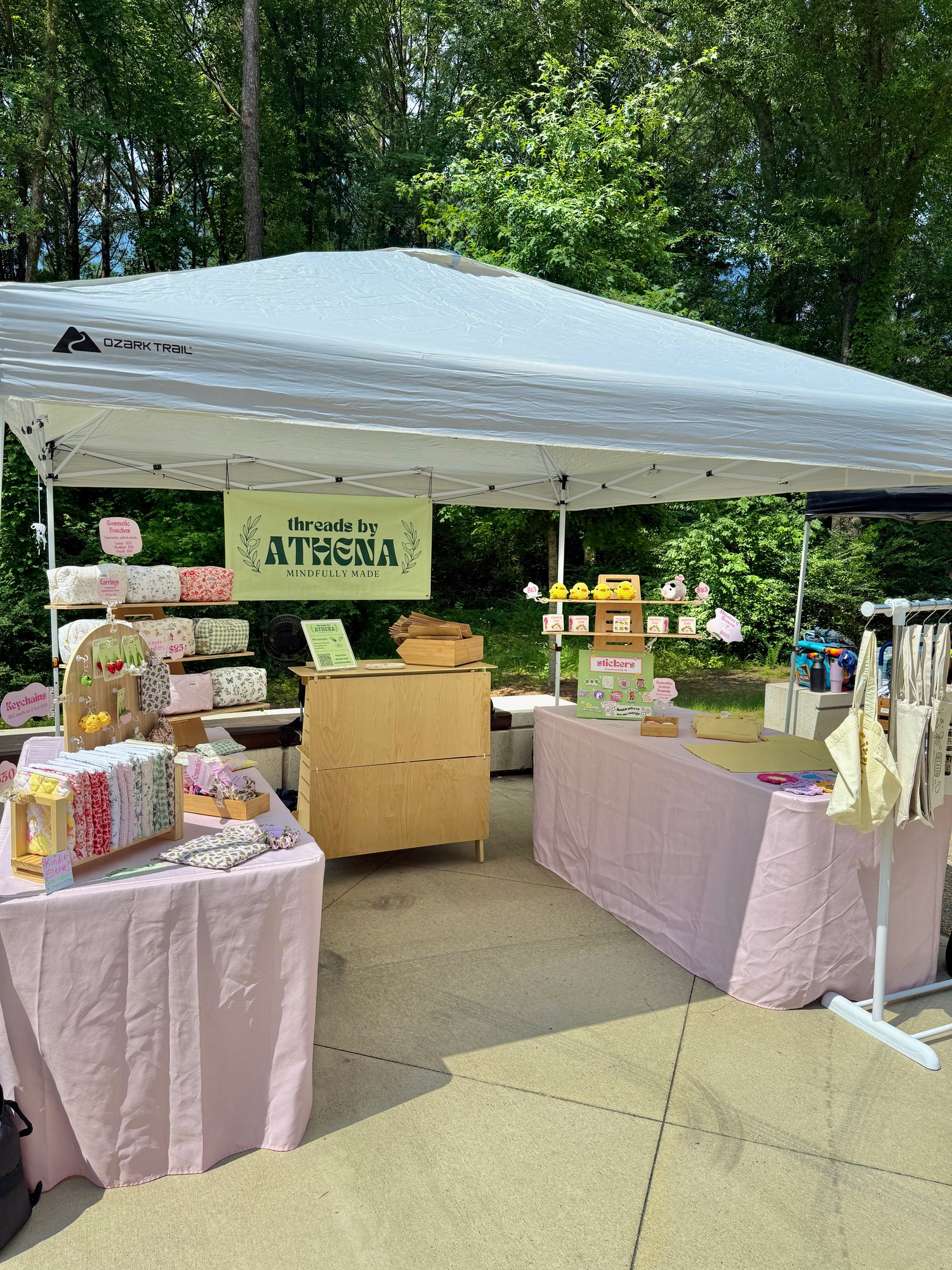 Outdoor market stall with various products under a white tent, surrounded by trees.