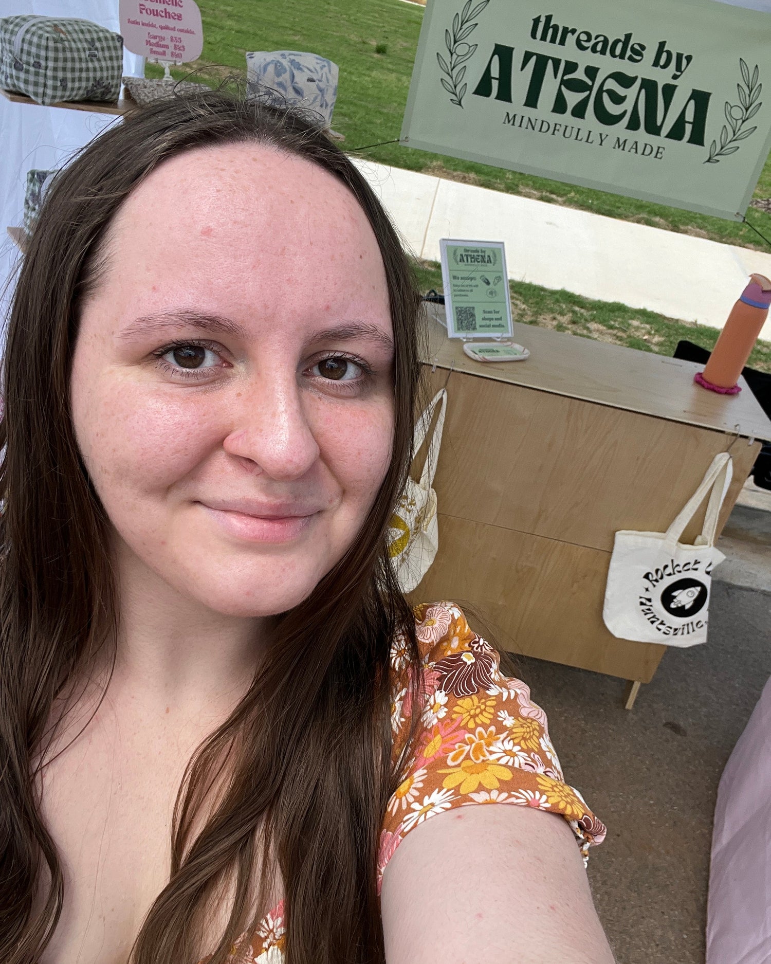 Person with long brown hair wearing a floral dress, standing in front of a wooden kiosk with 'threads by Athena' branding.
